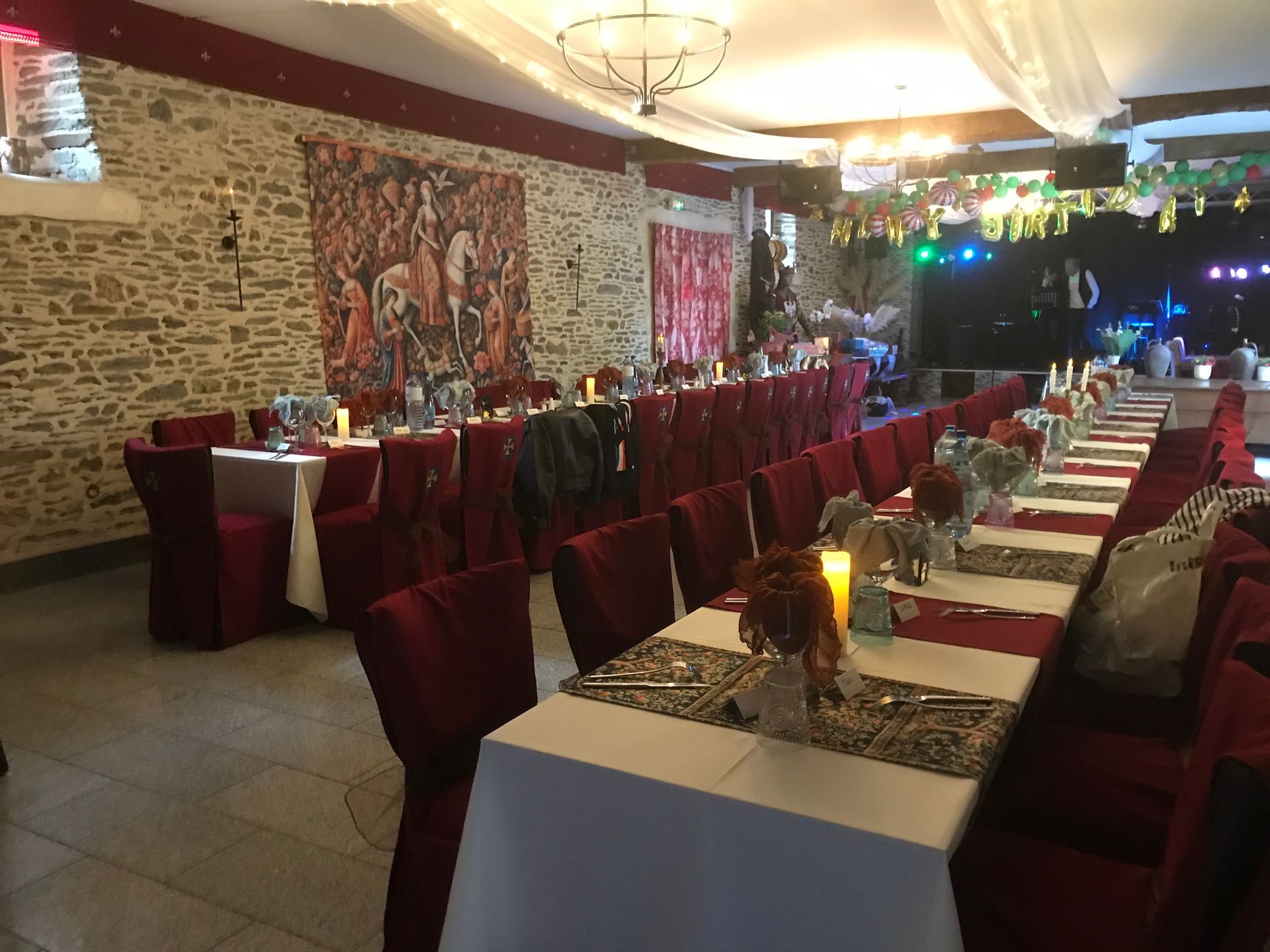 Une longue salle aménagée pour un événement formel, avec des tables nappées de blanc, des chaises rouges et des murs en pierre ornés d'une tapisserie.
