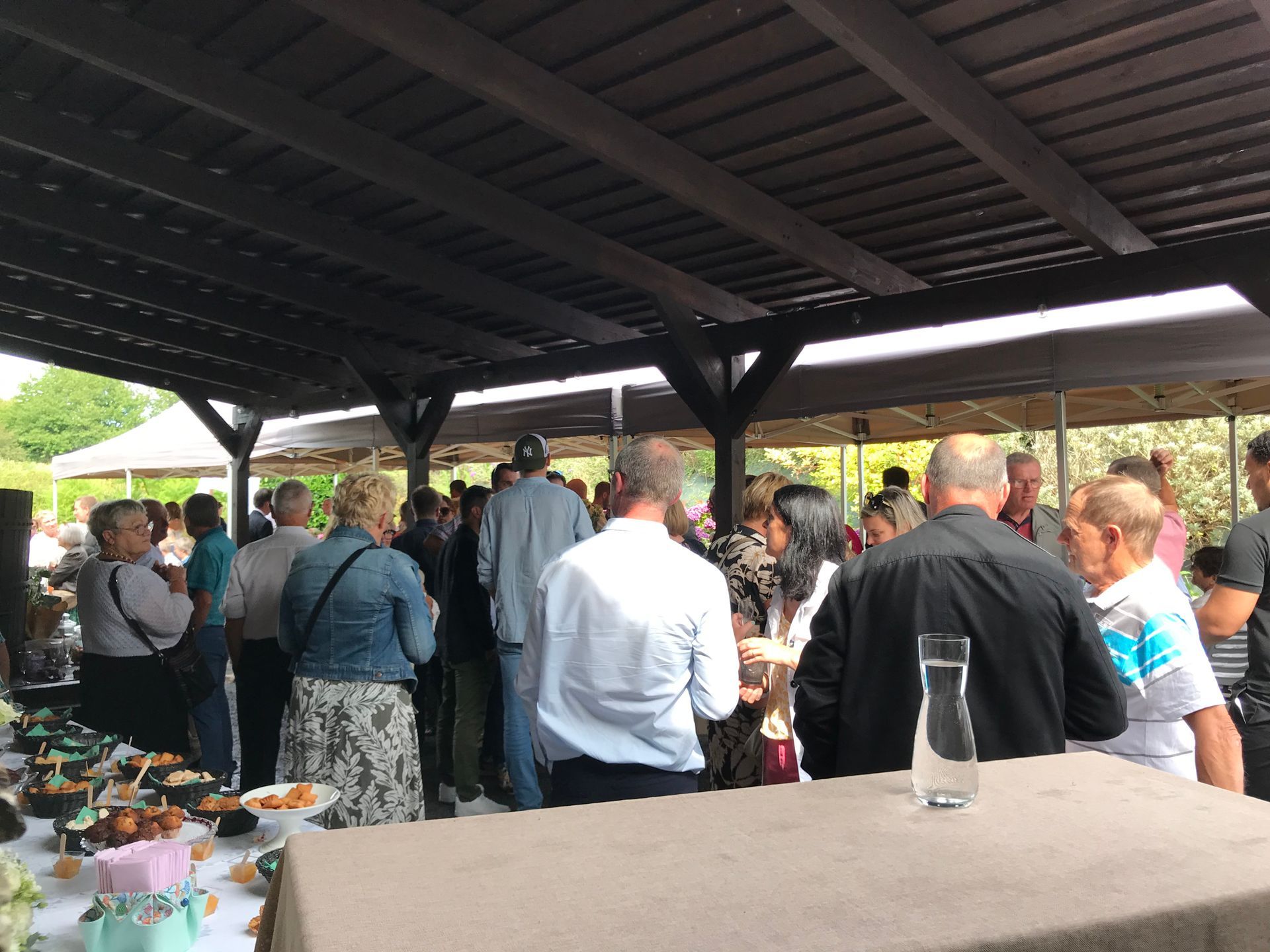 Un groupe de personnes se réunit pour un événement social en plein air sous une structure au toit en bois aménagée avec des tables et des en-cas.