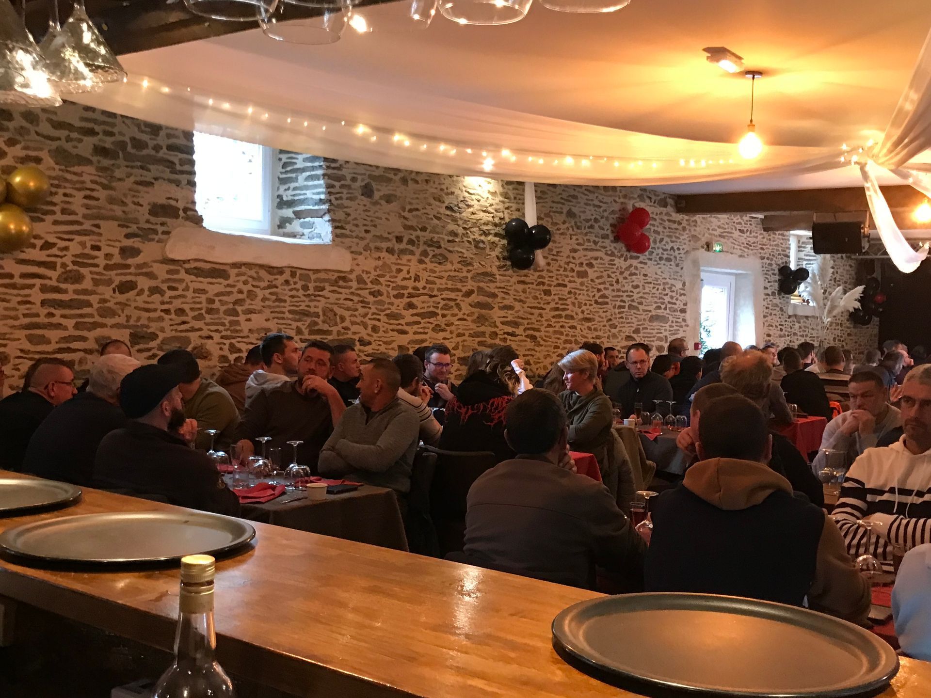 Un espace événementiel bondé, avec des murs en pierre rustiques, des guirlandes lumineuses et un bar en bois au premier plan, avec des plateaux vides.