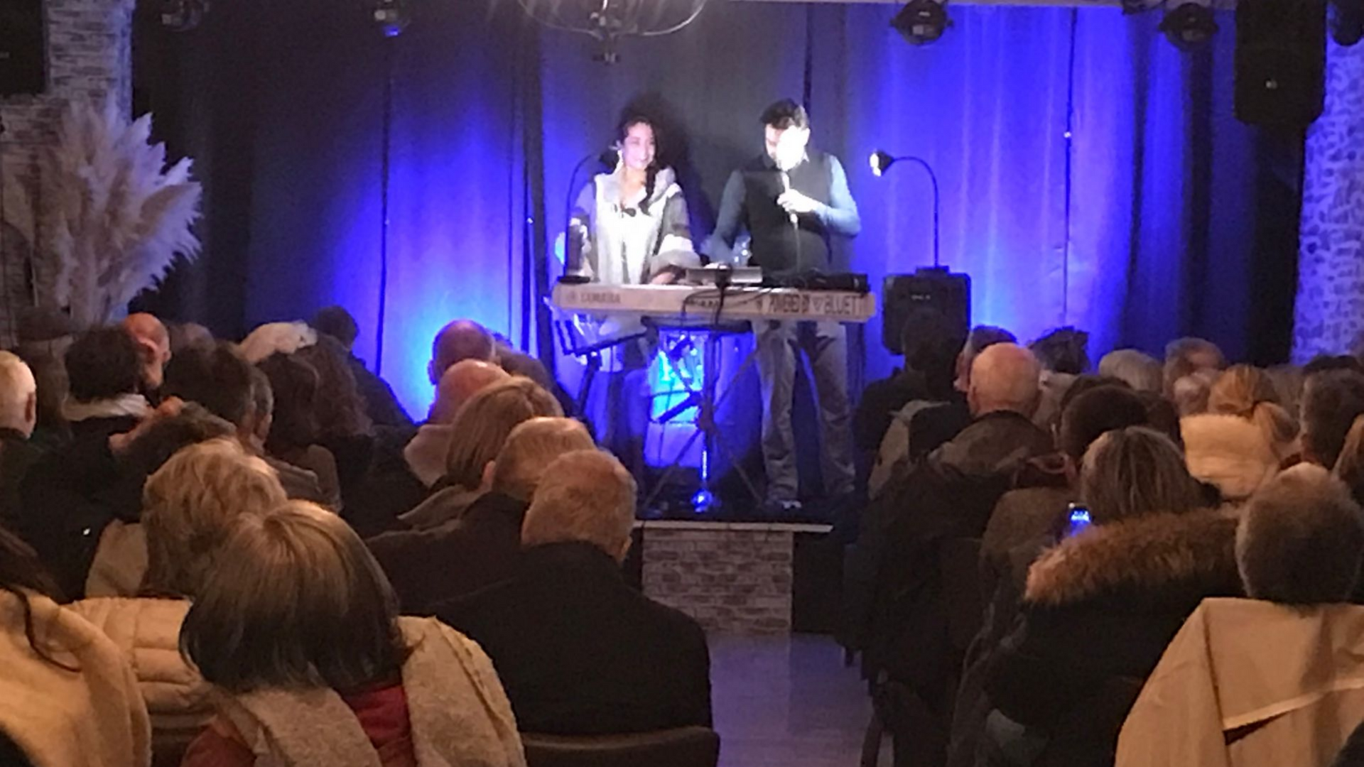 Deux personnes se produisent sur une scène éclairée de bleu devant un public assis dans une salle intérieure faiblement éclairée.