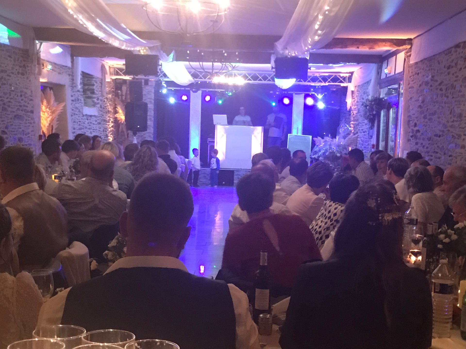 Une salle de banquet faiblement éclairée, avec des invités attablés face à une cabine de DJ illuminée par des projecteurs colorés.
