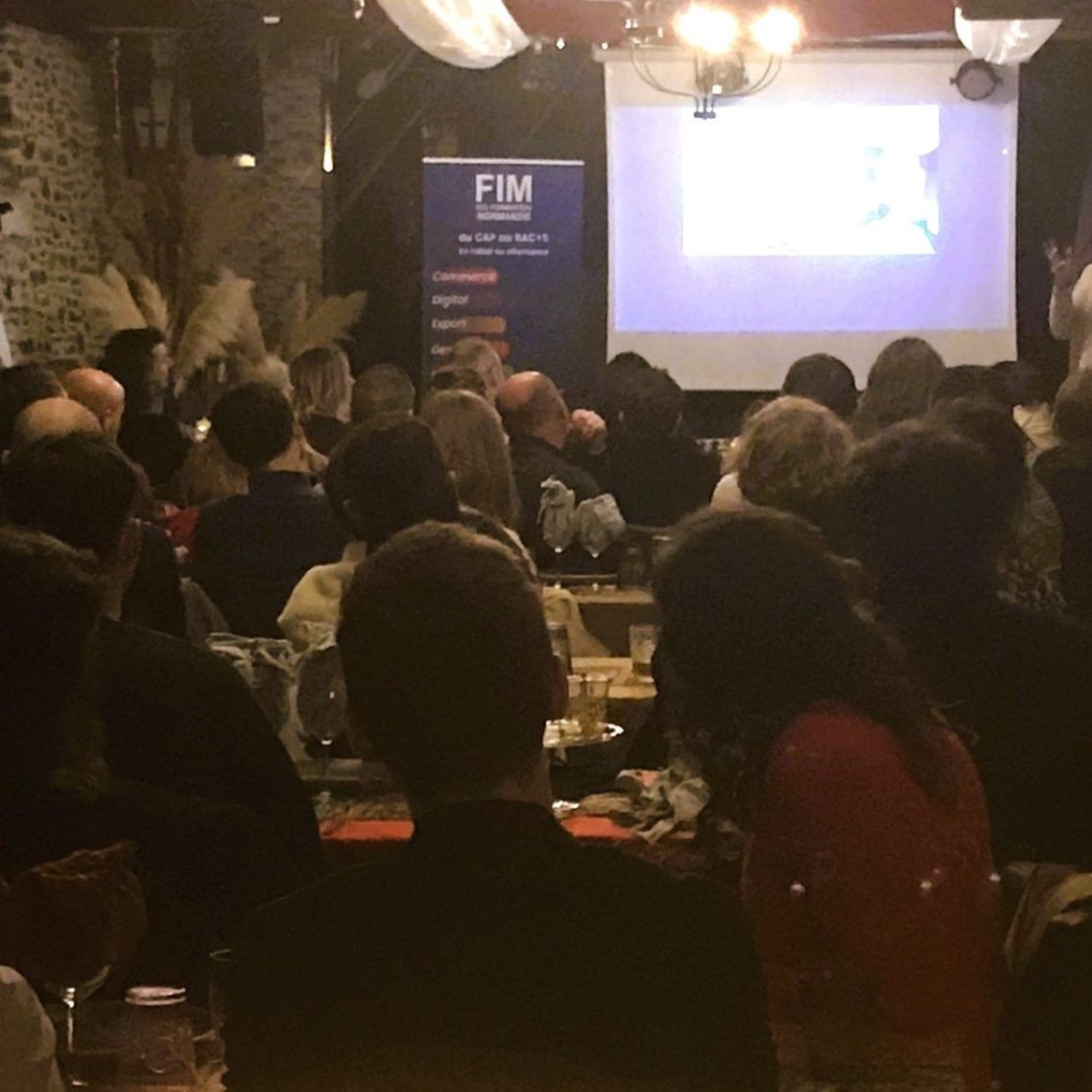 Un public assiste à une présentation dans une salle rustique faiblement éclairée, équipée d'un écran de projection.