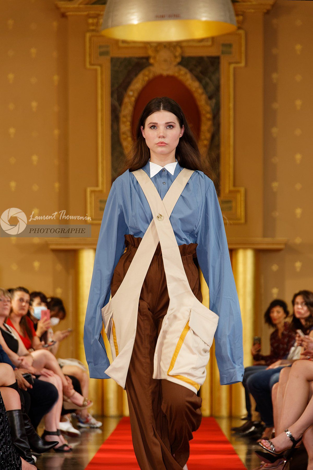 Mannequin défilant sur le tapis rouge, vêtue d'une chemise bleue, d'un pantalon marron et d'un pantalon beige à poches façon plastron. Spectateurs.