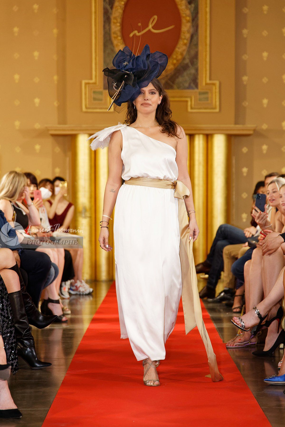 Mannequin en robe blanche asymétrique et chapeau bleu marine défilant sur le tapis rouge, accents dorés, public.
