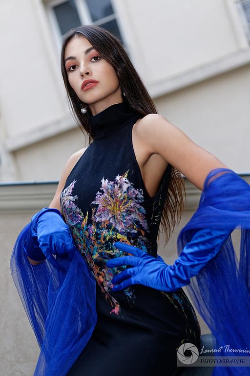 Femme en robe noire à broderies florales et gants bleus, debout à l'extérieur.