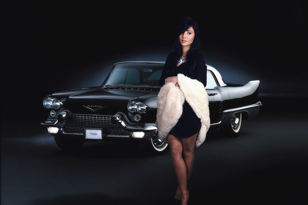 Une femme en robe noire et châle de fourrure se tient devant une voiture de collection noire.