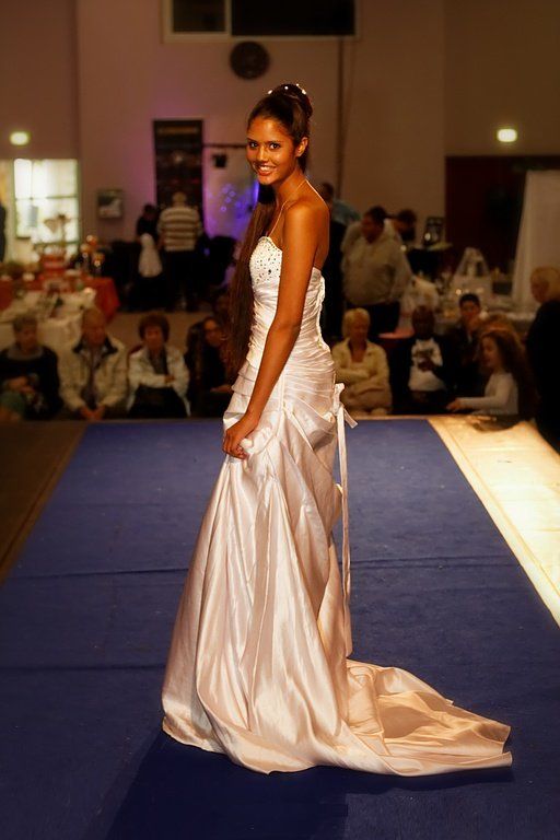 Une femme en robe blanche bustier pose sur un podium bleu lors d'un défilé de mode, souriante.
