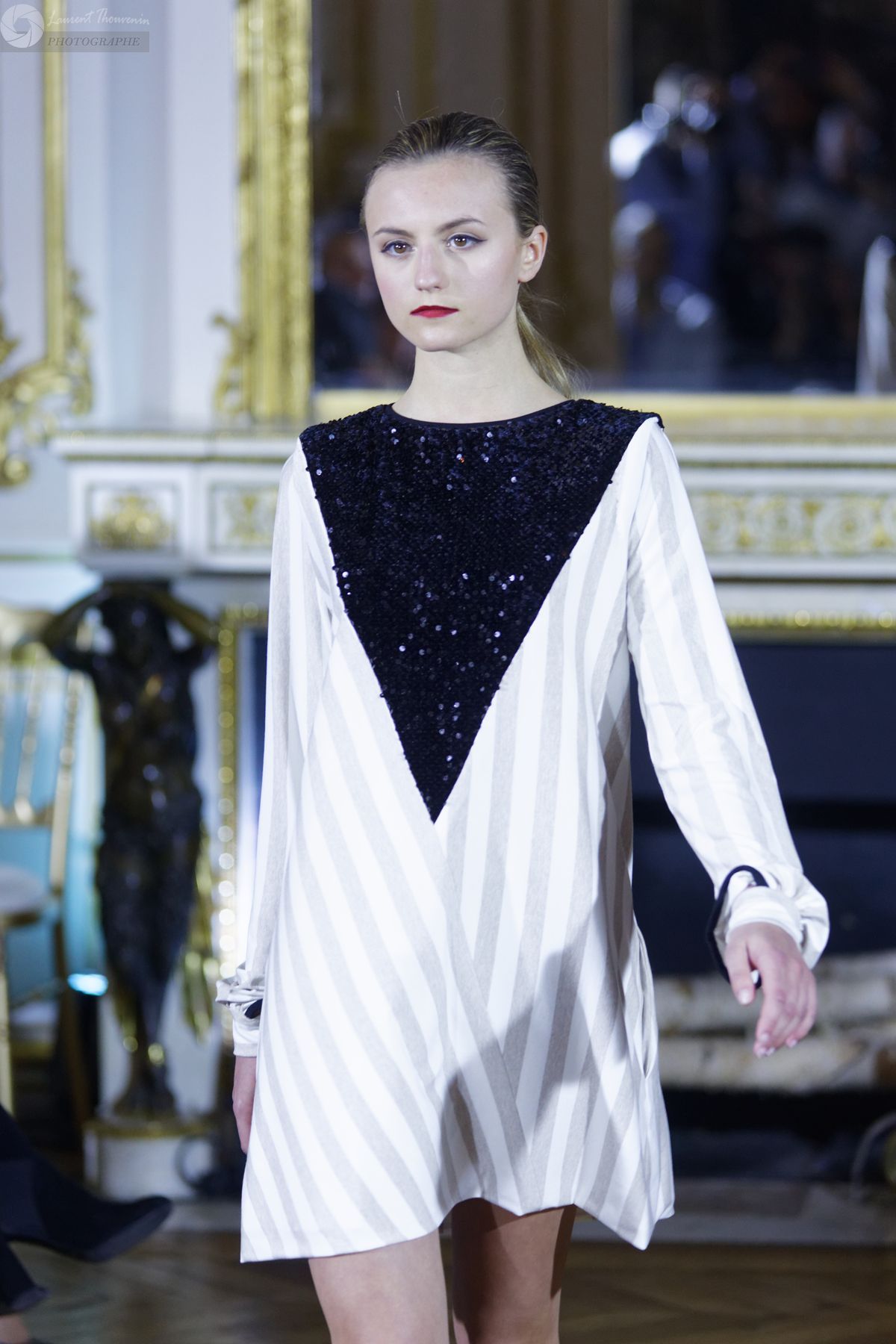 Mannequin portant une robe à rayures blanches avec un corsage à paillettes noires, défilant sur un podium.