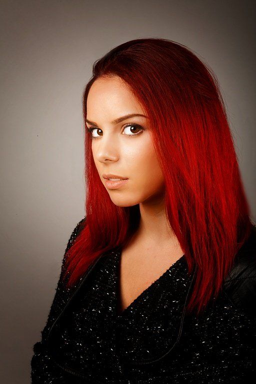 Femme aux cheveux roux flamboyants regardant l'objectif, vêtue d'un haut noir sur fond gris.