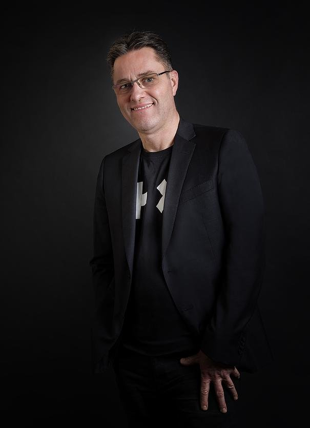 Un homme portant des lunettes, une veste noire et un t-shirt, souriant, les mains dans les poches, se détache sur un fond sombre.