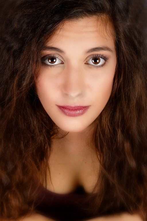 Femme aux longs cheveux bruns et bouclés, regardant droit dans l'objectif, les yeux foncés, les lèvres foncées et le décolleté plongeant.