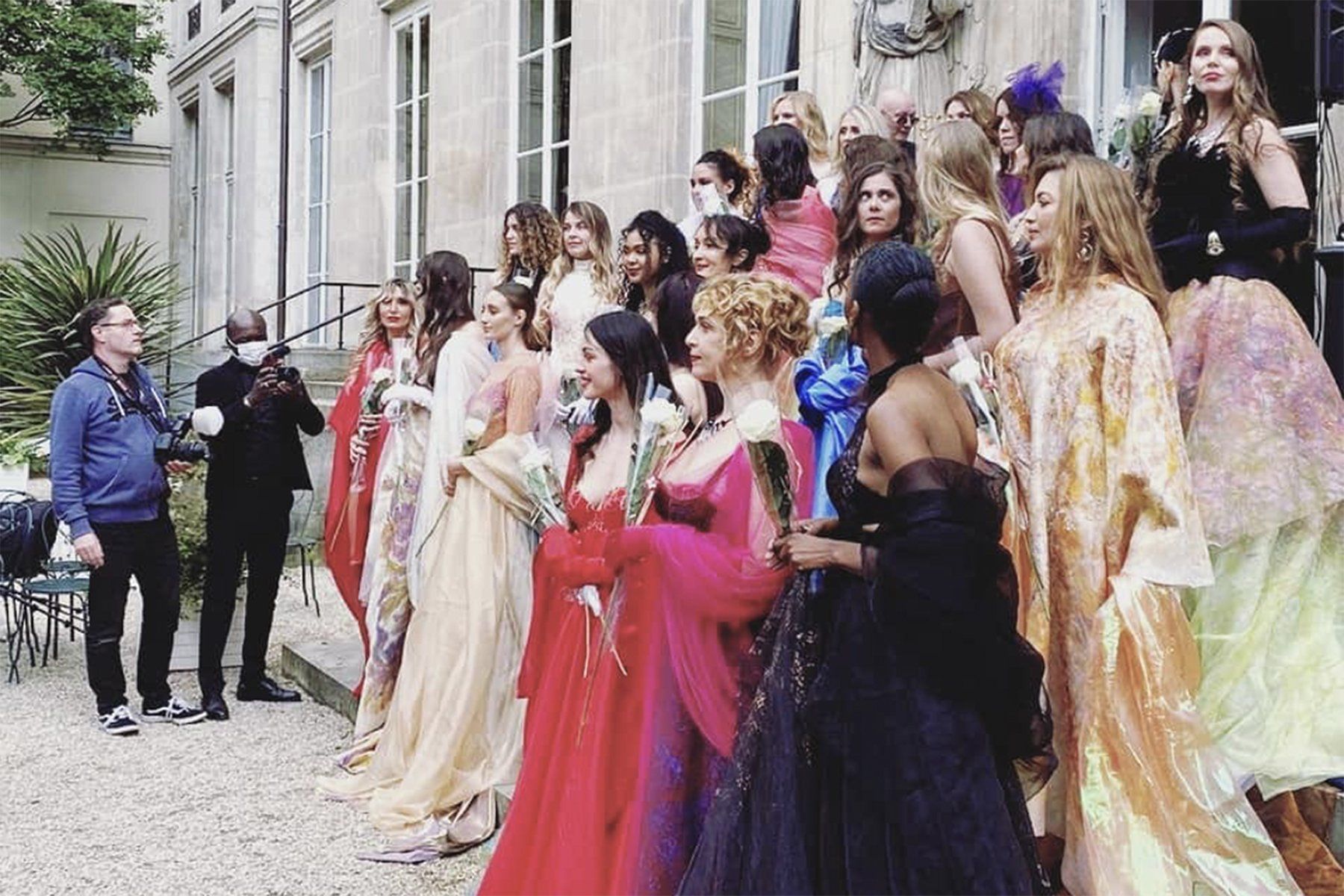 Un groupe de personnes en robes pose sur les marches d'un immeuble. Le photographe est visible en arrière-plan.
