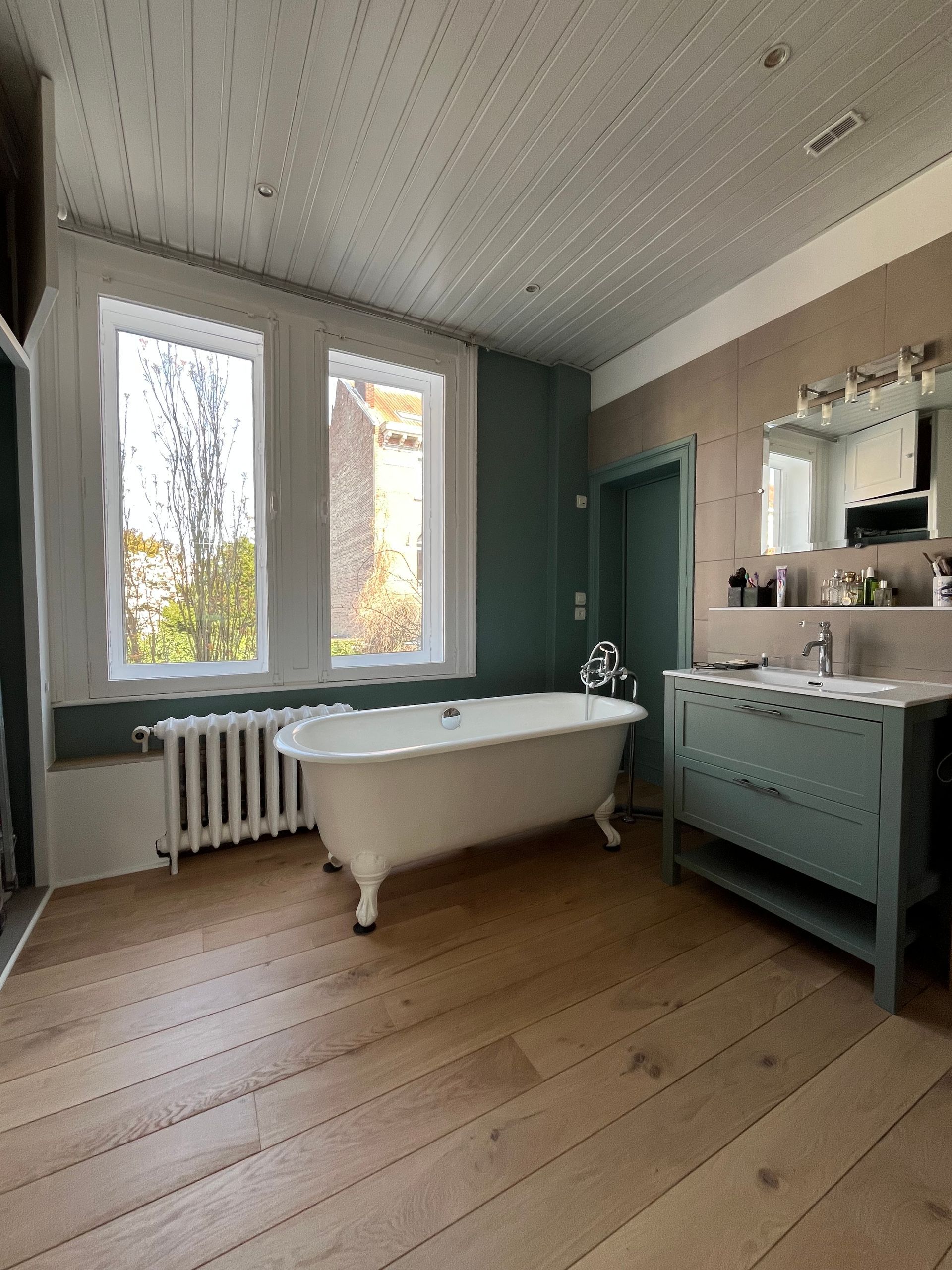Salle de bains avec baignoire sur pieds, deux fenêtres, parquet et touches de couleur turquoise.