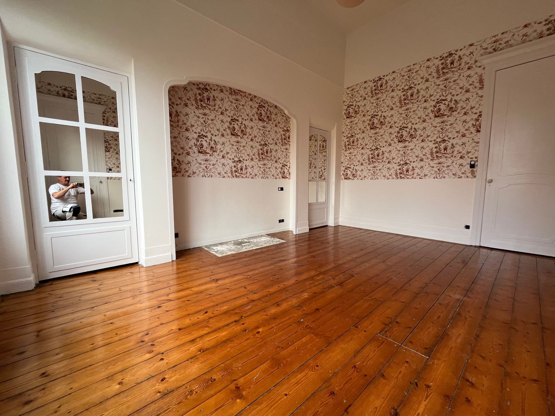 Pièce avec parquet, murs et moulures blancs, partiellement tapissée ; une personne peint une fenêtre.