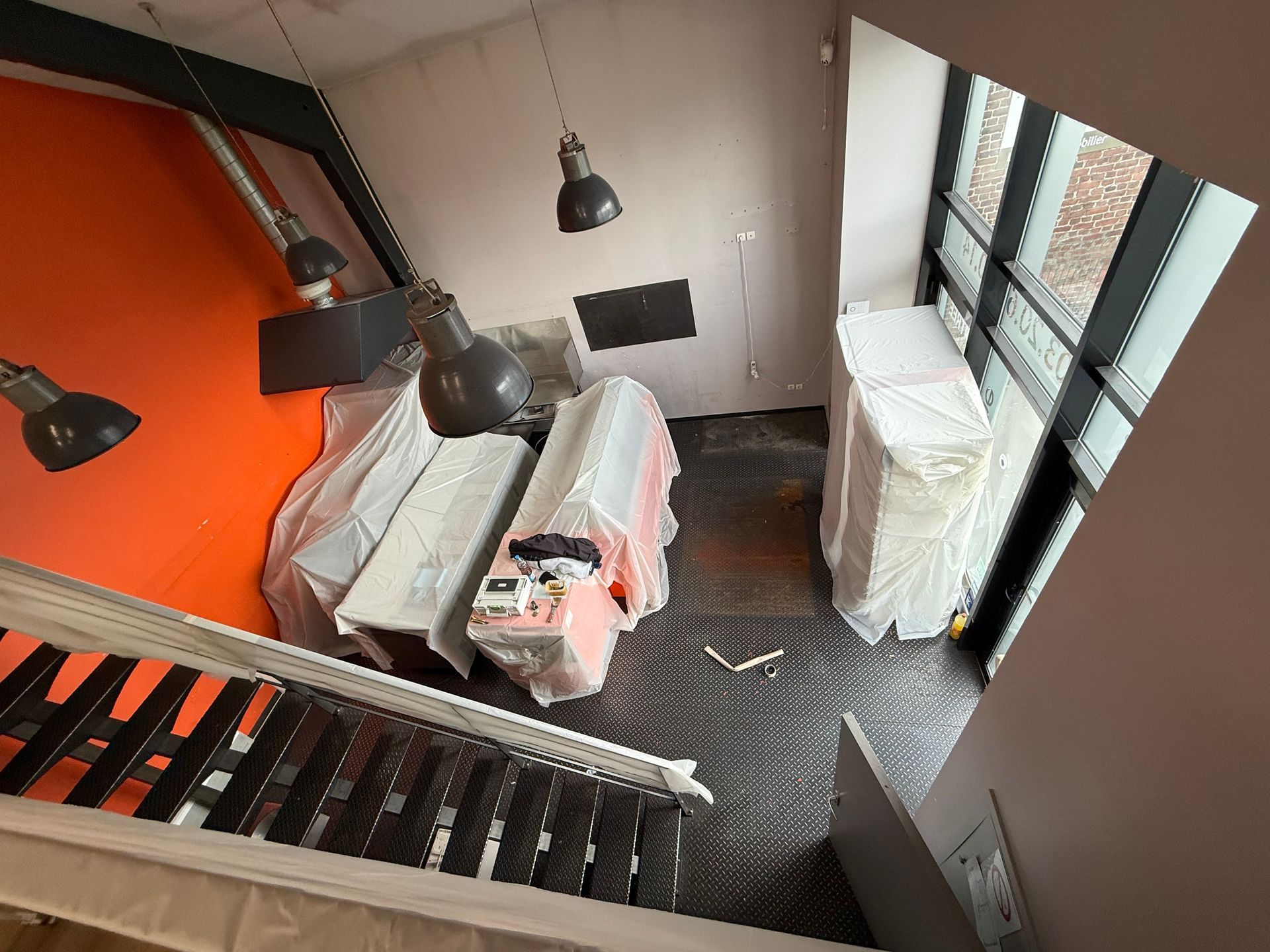 Pièce intérieure avec meubles recouverts, mur orange, escalier et éclairage industriel.