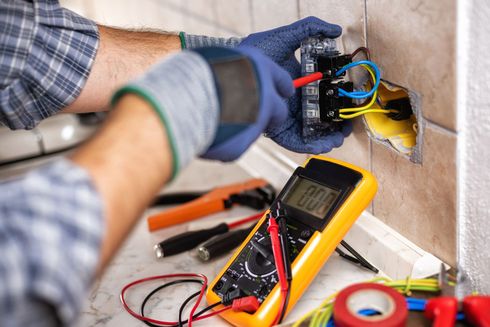 Un électricien portant des gants bleus installe une prise de courant dans un mur carrelé, avec un multimètre.