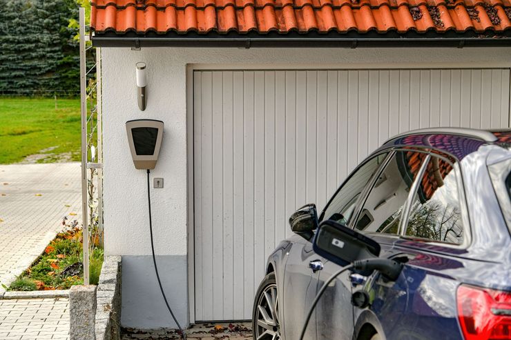 Une voiture sombre garée devant une porte de garage blanche, branchée à une borne de recharge fixée au mur extérieur.