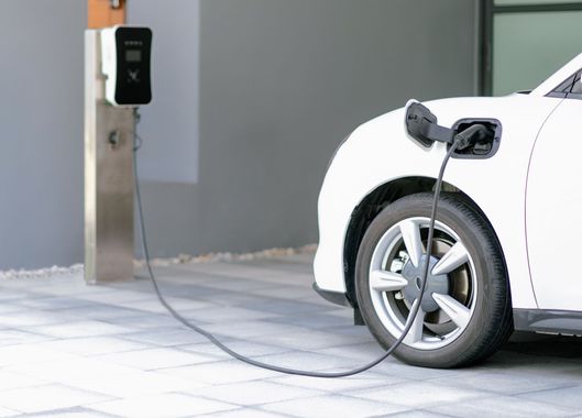 Une voiture électrique blanche garée sur une allée en pierre, branchée à une borne de recharge murale.