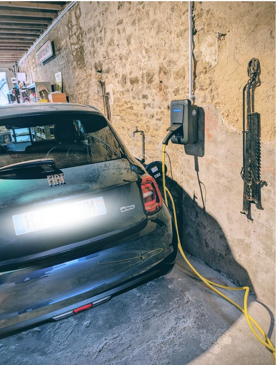 Une Fiat 500 électrique grise branchée à une borne de recharge murale à l'intérieur d'un garage aux murs de pierre.