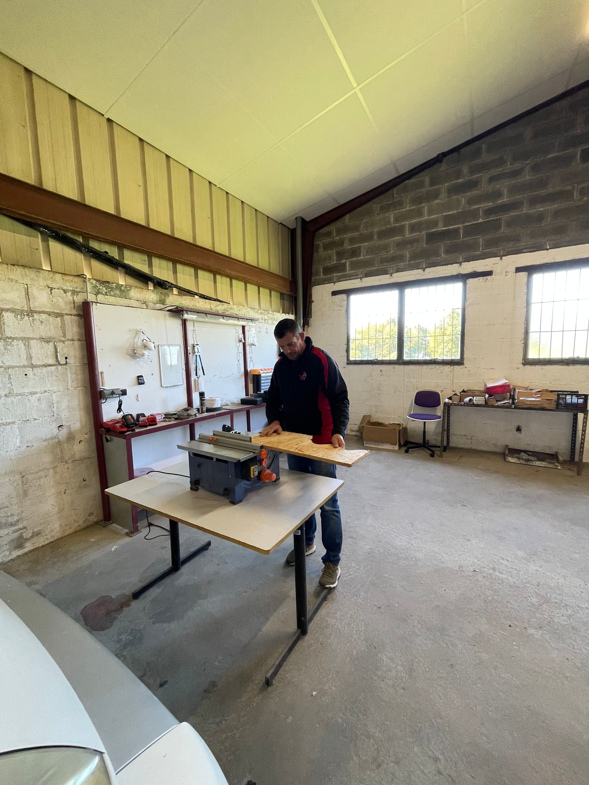 Homme utilisant une scie circulaire pour couper du bois dans un atelier. Murs et fenêtres gris.