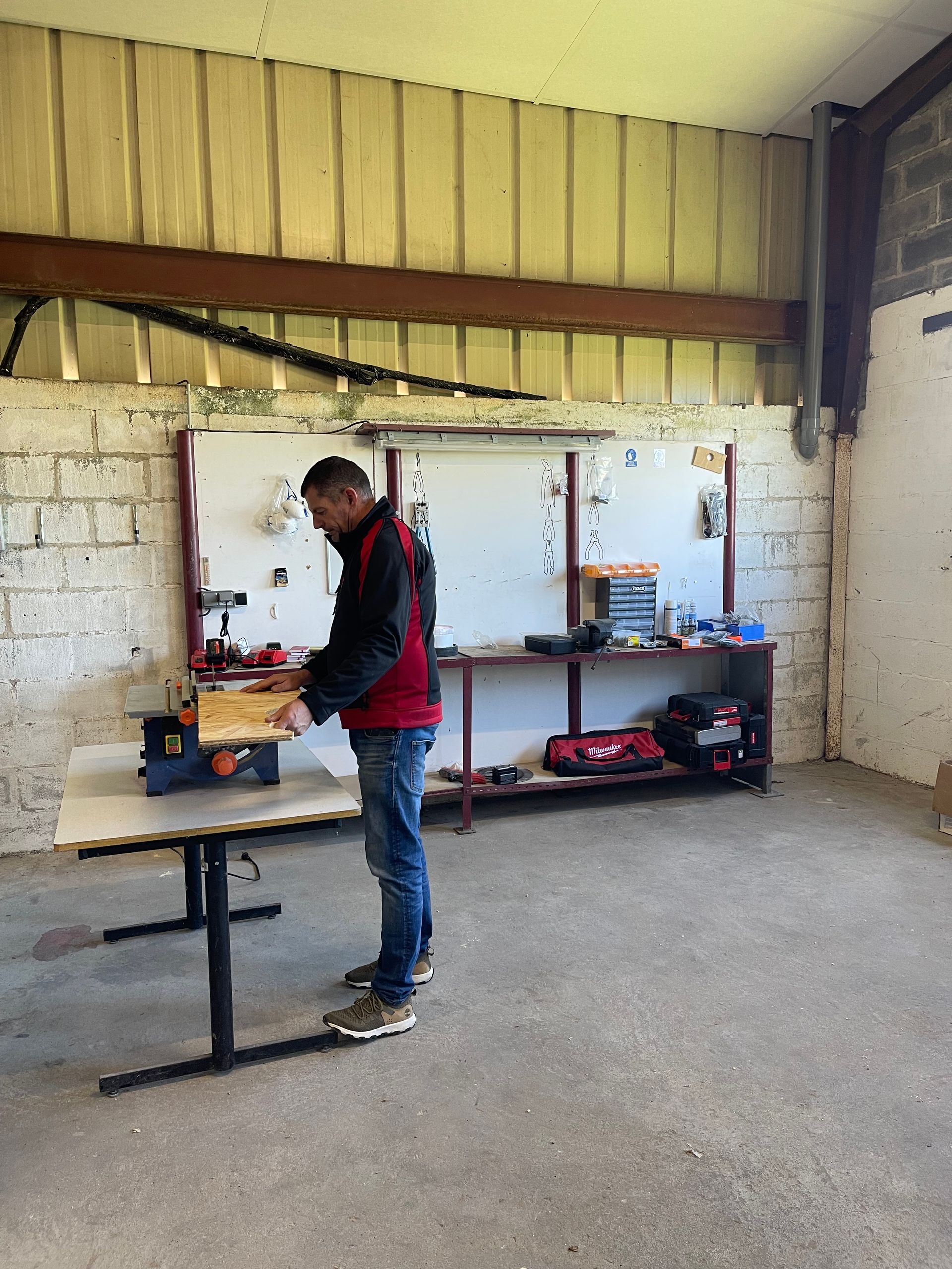 Homme travaillant à un établi dans un atelier. Veste rouge et noire, jean, outils sur des étagères.