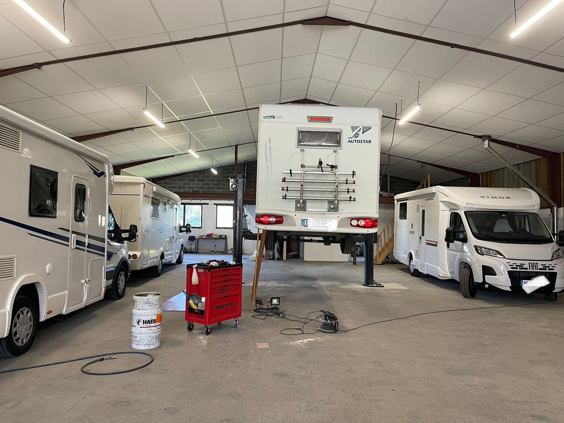 Camping-cars dans un atelier. L'un d'eux est surélevé pour les réparations ; une boîte à outils se trouve à proximité.