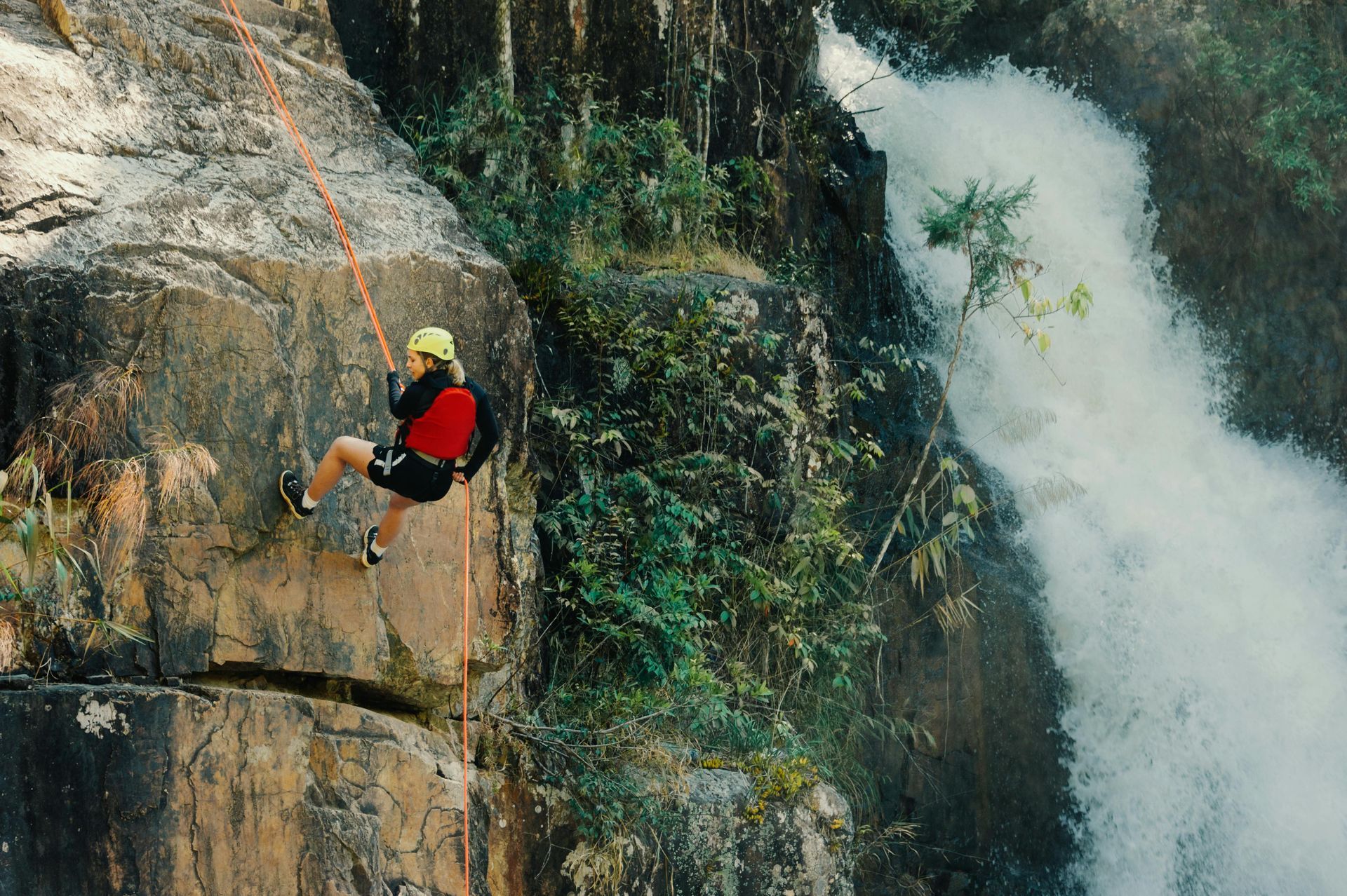 Man performing extreme rock climbing with by a large waterfall and trees 