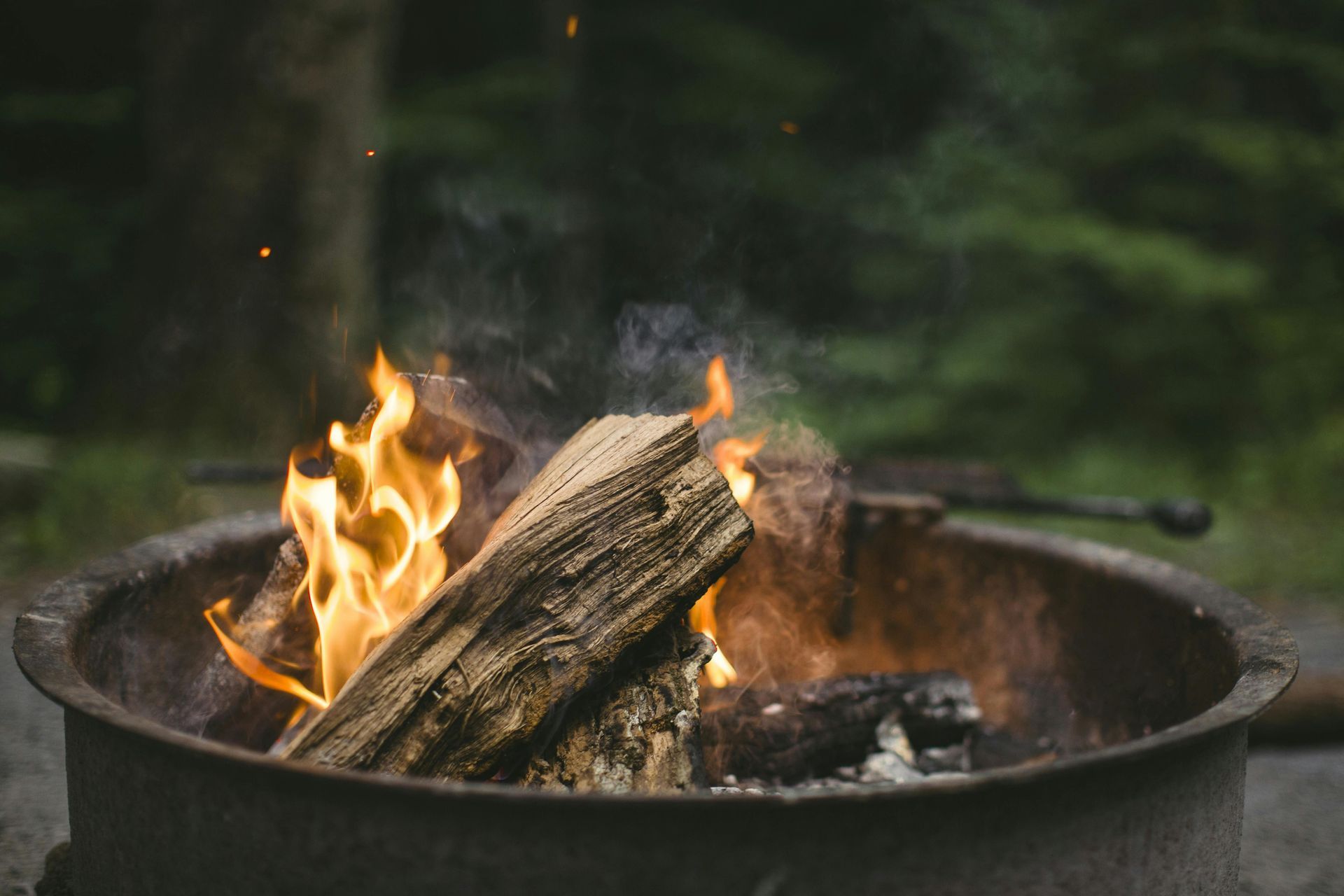 Active campfire in metal firepit outdoors with blurred green trees in background