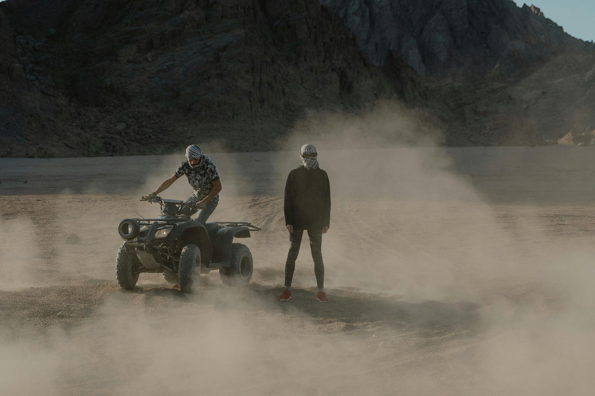 Person riding ATV in a field, kicking up dirt around another person 