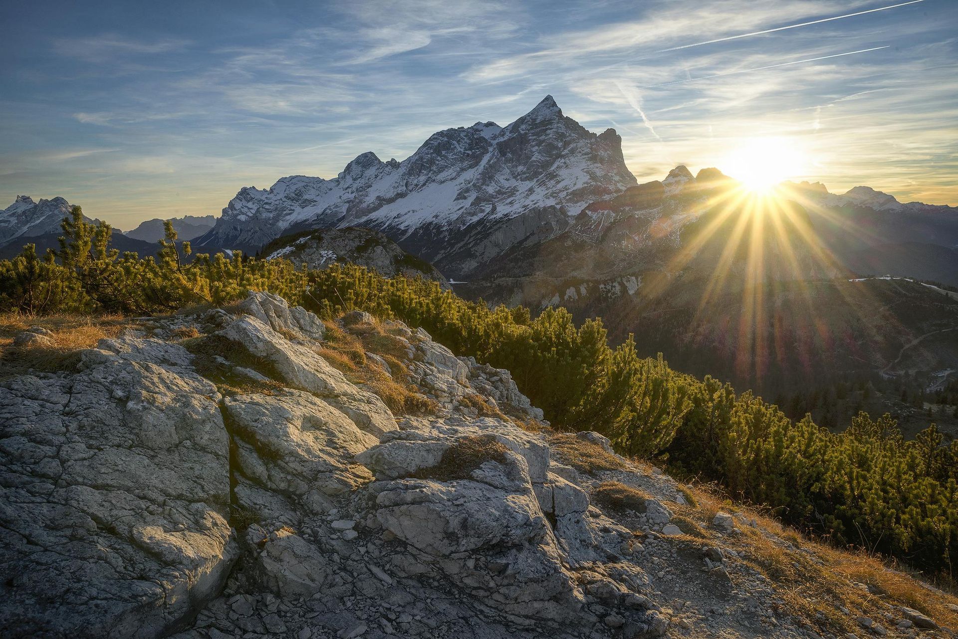 Sun peaking over mountains with some snow on top and sunlight hitting the rocks