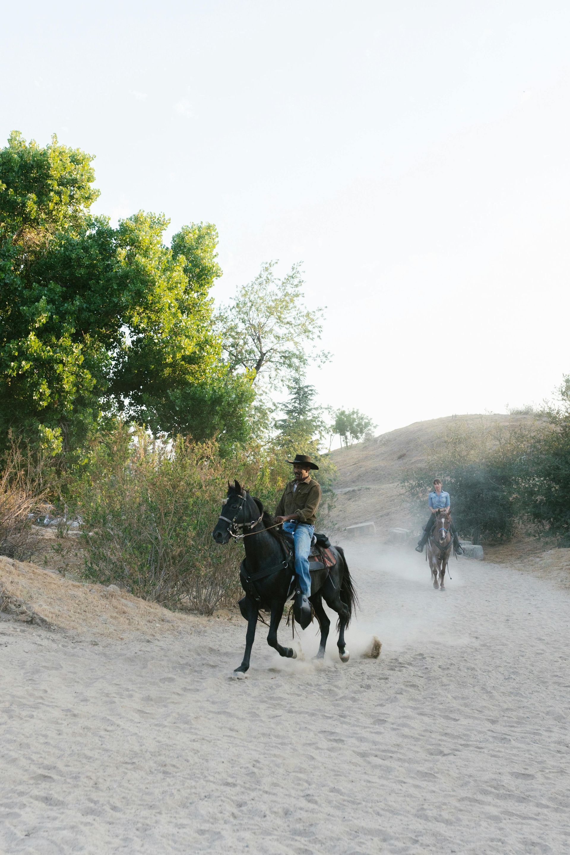 Two people riding horses throughout dirt trail on a bright, clear sky day