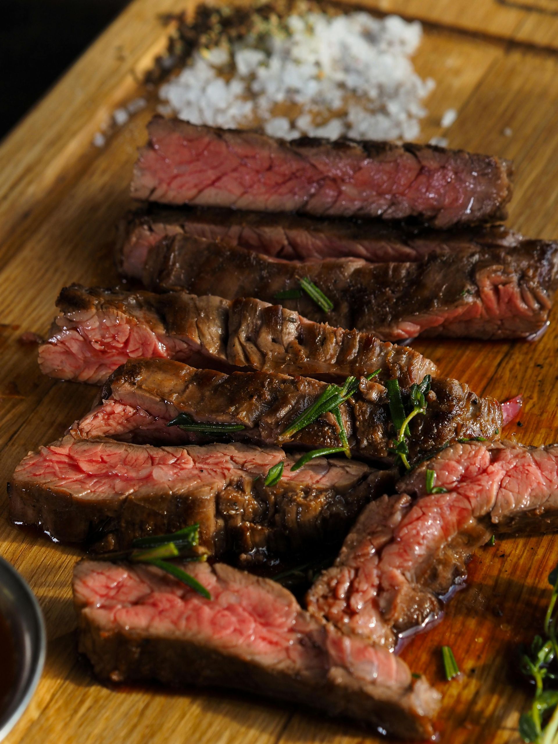 Sliced medium-rare steak on a wooden board with rosemary and sauce.