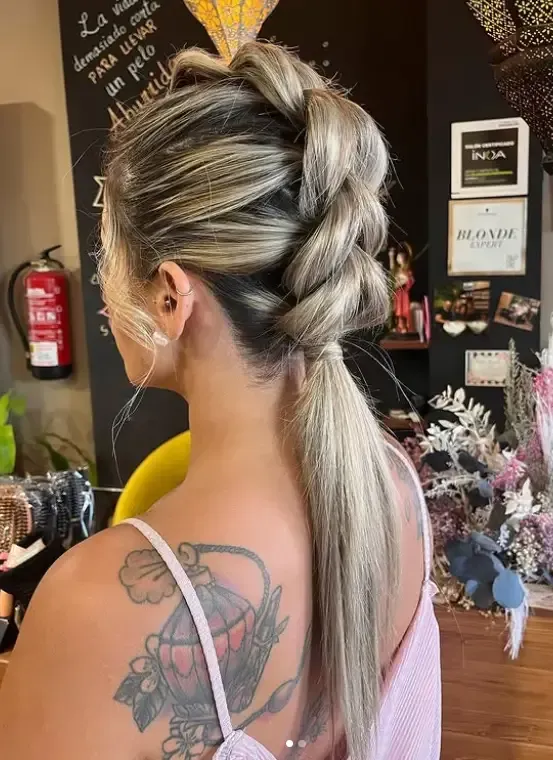 Vista trasera de una persona con cabello rubio con mechas, peinado en una cresta trenzada que termina en una coleta baja.