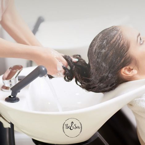 Una persona recibe un relajante lavado de cabello en un salón de belleza, con espuma jabonosa en la cabeza y productos tipo spa sobre una mesa de madera.