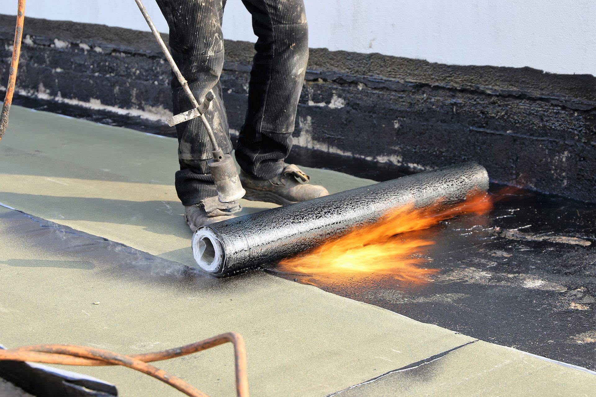 Ein Arbeiter brennt eine ausgerollte Dachmembran auf einem Flachdach ab; Flammen und Rauch sind sichtbar.
