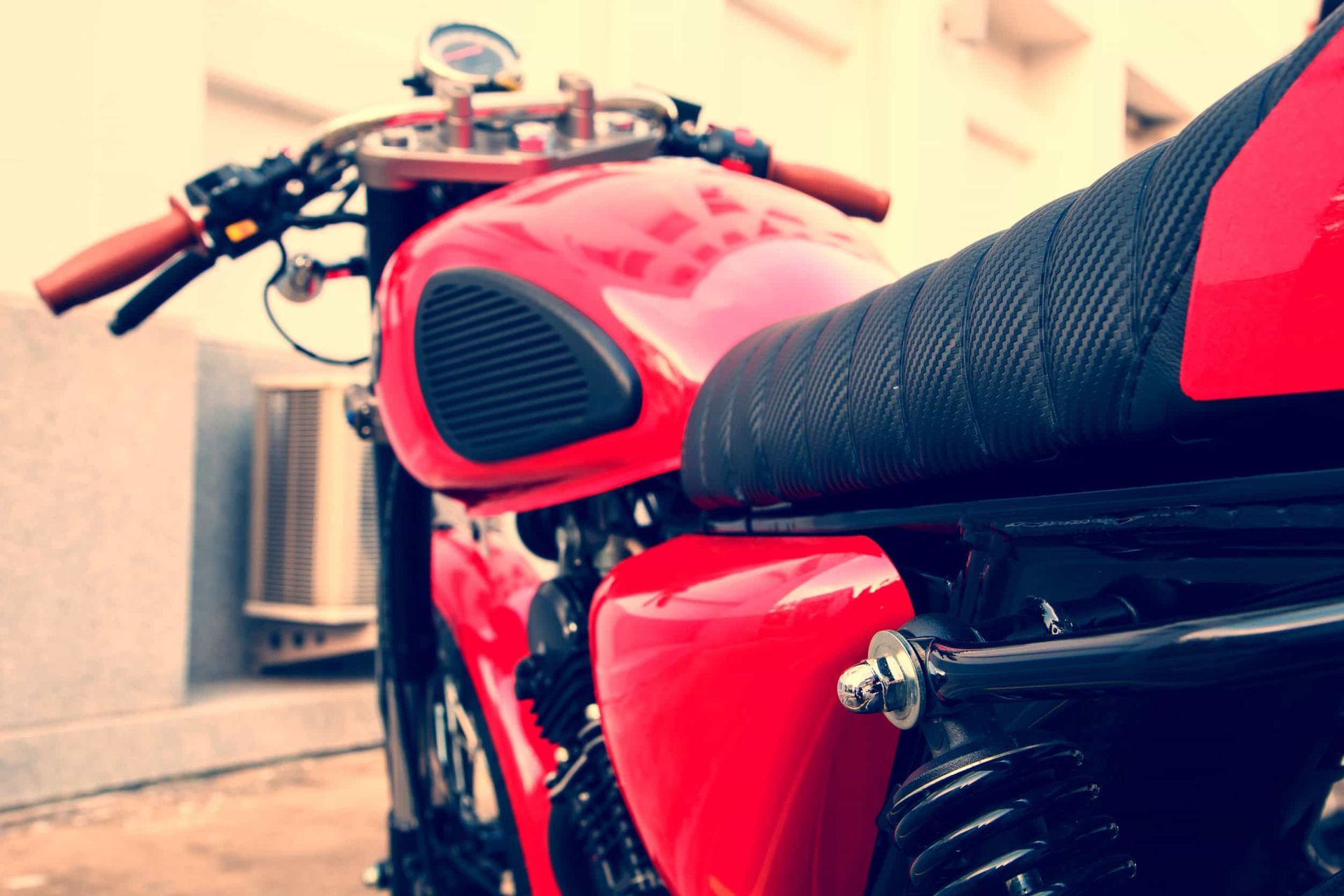 Primer plano de una motocicleta roja con el depósito brillante y el asiento negro, aparcada al aire libre junto a un edificio.