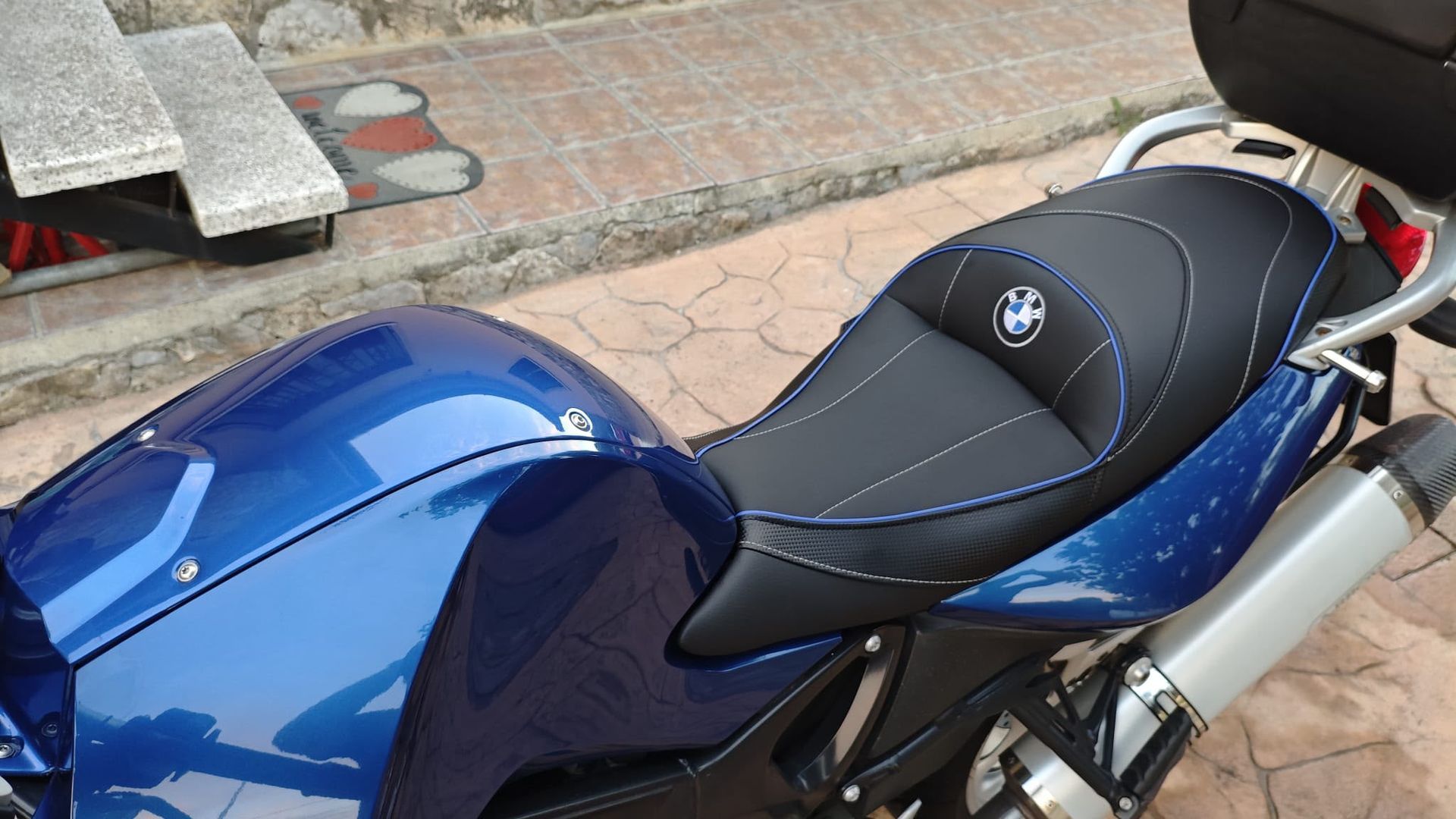 Asiento y depósito de combustible azules de una motocicleta BMW aparcada al aire libre sobre una acera de ladrillo.