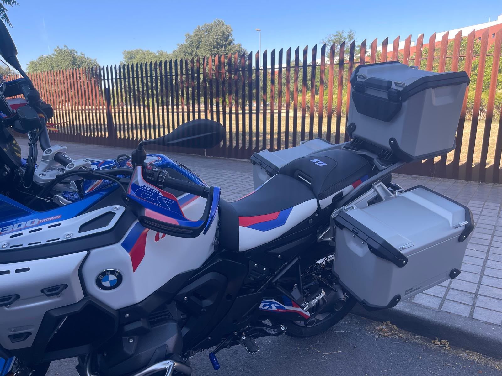 Motocicleta BMW touring plateada y azul aparcada junto a una valla, con baúl trasero y maletas laterales.