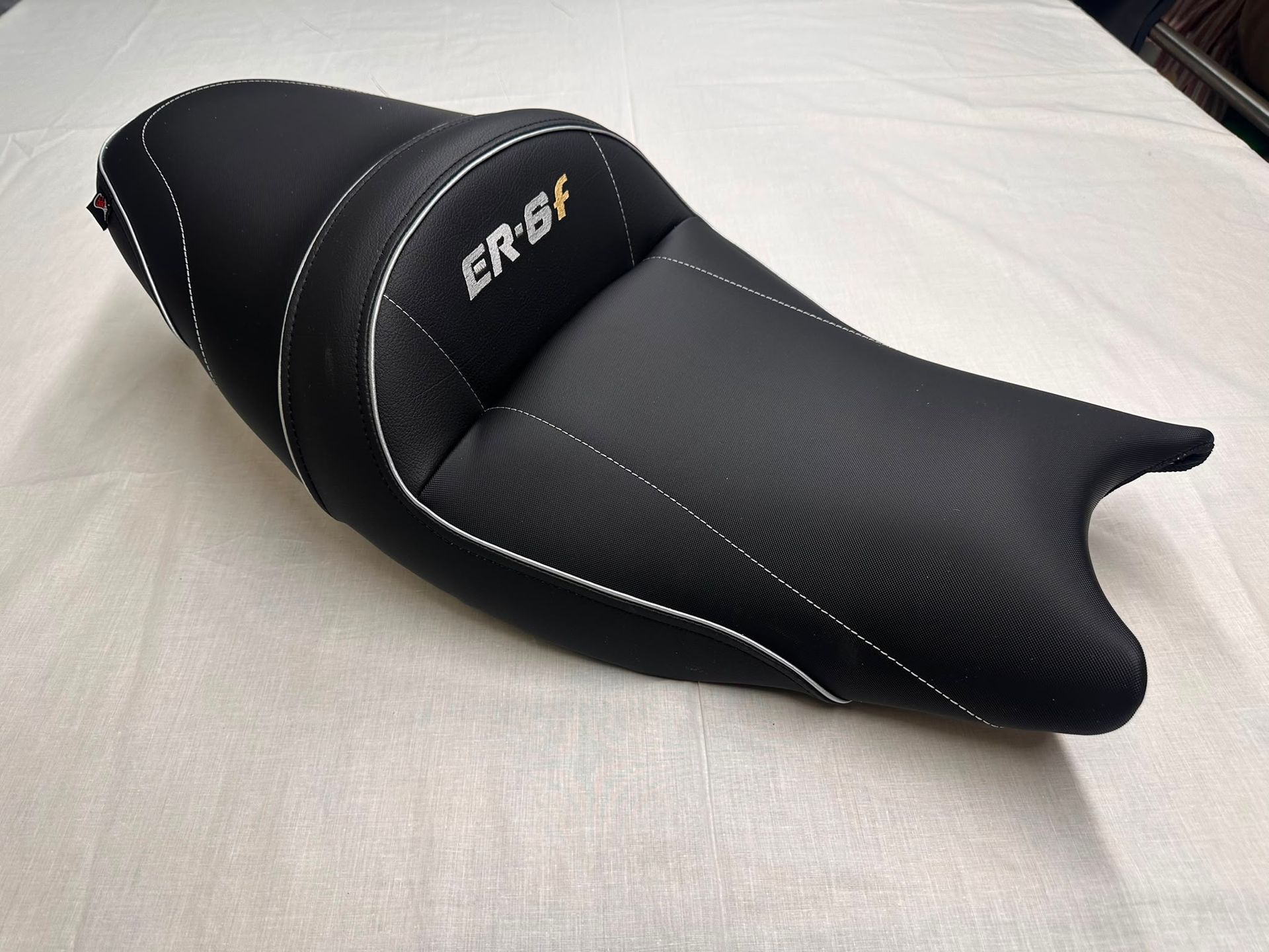 Asiento negro para motocicleta con costuras blancas, sobre un fondo liso y claro