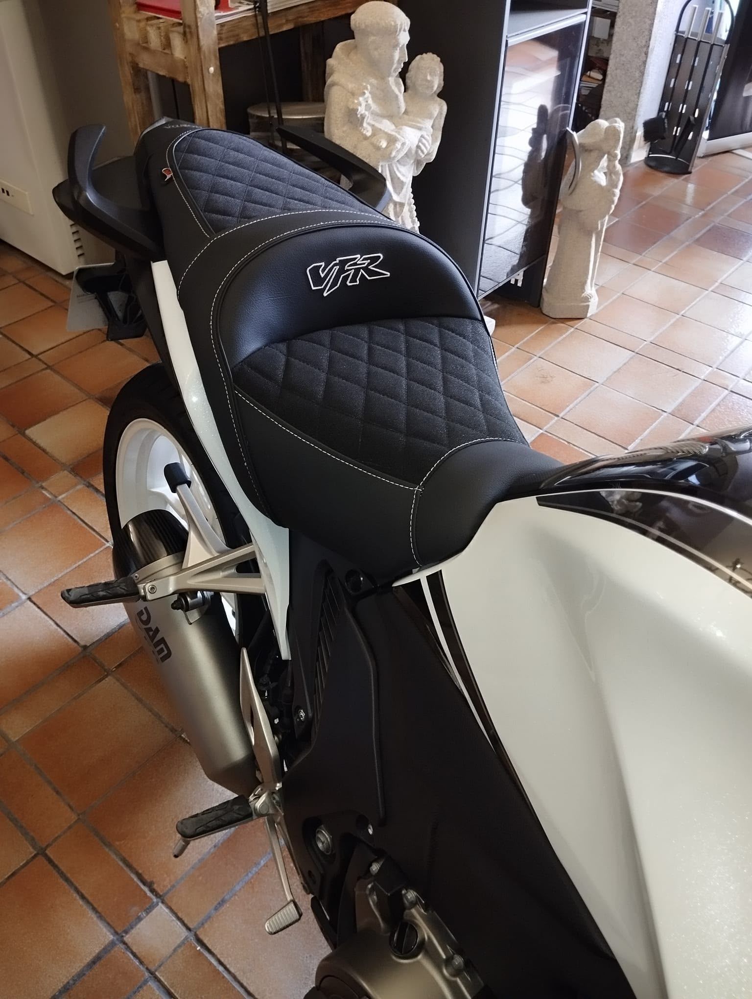 Asiento negro de motocicleta y carrocería blanca aparcados sobre un suelo de baldosas en el interior, cerca de una chimenea.