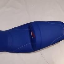 Un asiento de motocicleta de color azul brillante con finas costuras blancas, visto desde arriba sobre un fondo blanco.