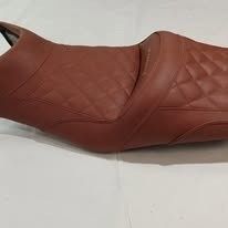 Un asiento de motocicleta marrón con costuras en forma de diamante y diseño escalonado para dos personas, que descansa sobre una superficie blanca.