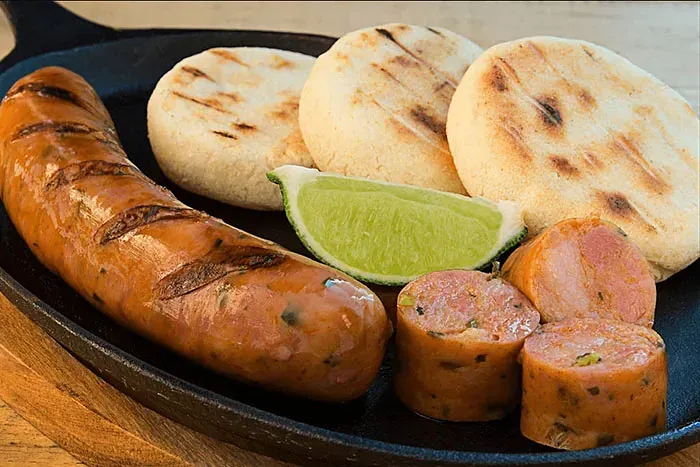 Una sartén llena de salchicha, arepas y una rodaja de limón.