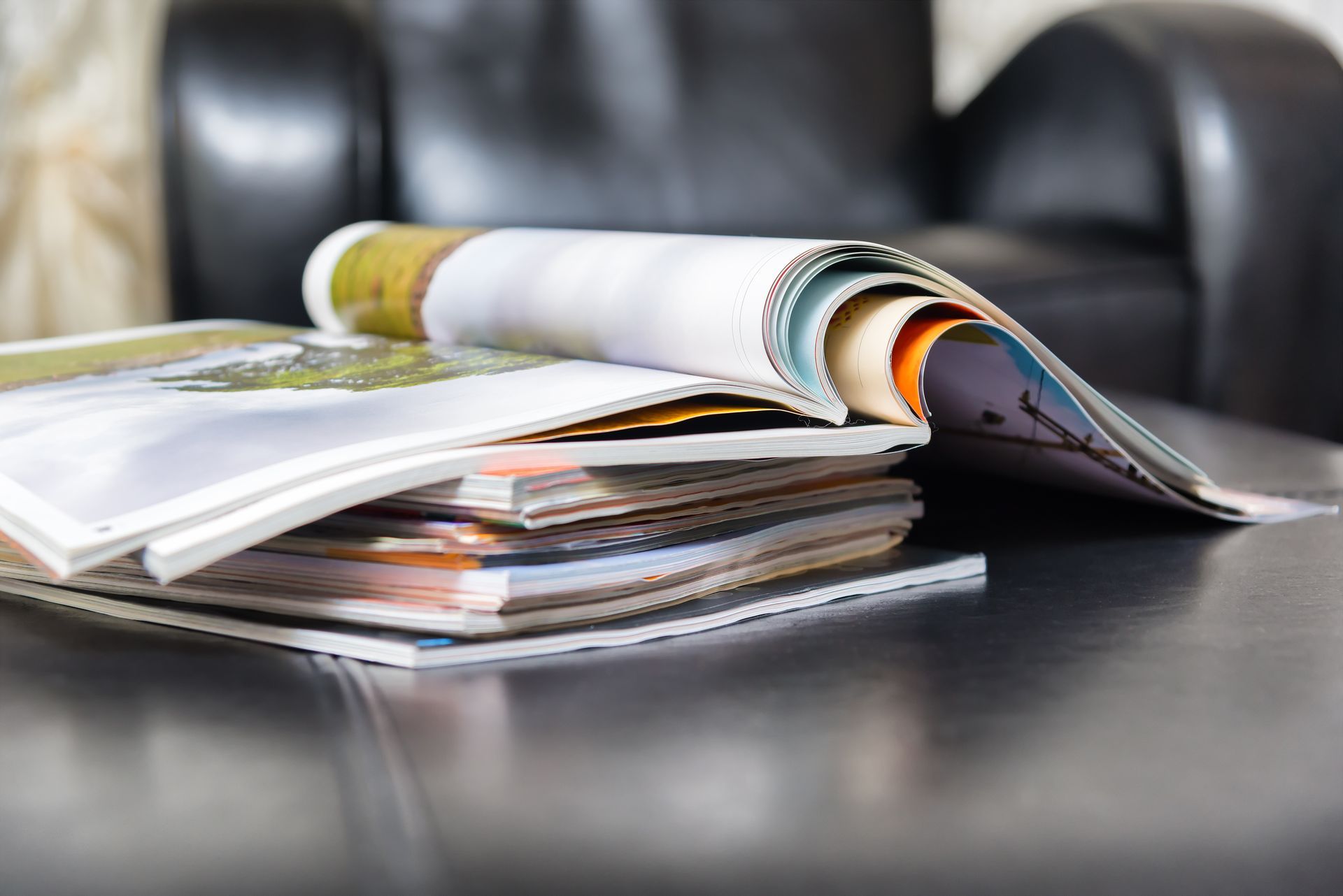 Une pile de magazines colorés est posée sur une surface en cuir noir devant un fauteuil à l'aspect flou.