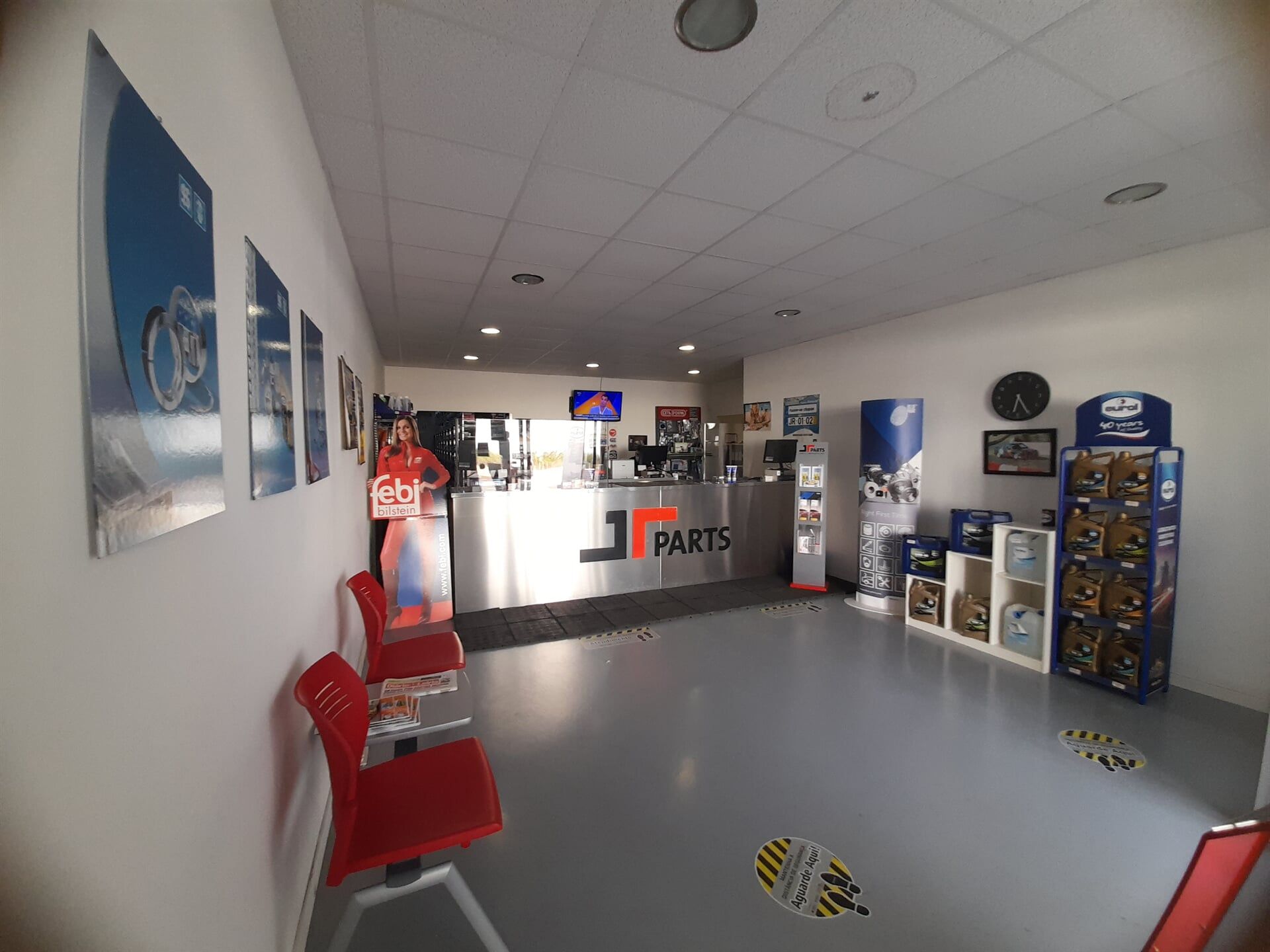 Interior de uma loja, com cadeiras, cartazes, um balcão e produtos em exposição. Uma pessoa está atrás do balcão.