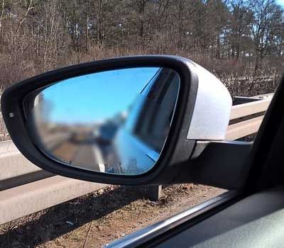 Retrovisor lateral mostrando um carro desfocado em uma estrada ao lado de guarda-corpos e árvores.