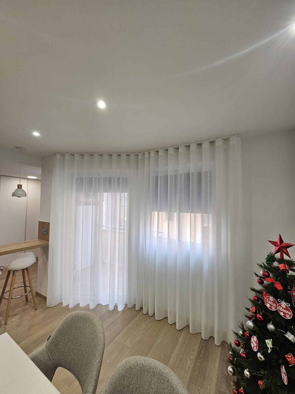 Comedor luminoso con mesa y sillas blancas, y cortinas transparentes sobre grandes ventanales.
