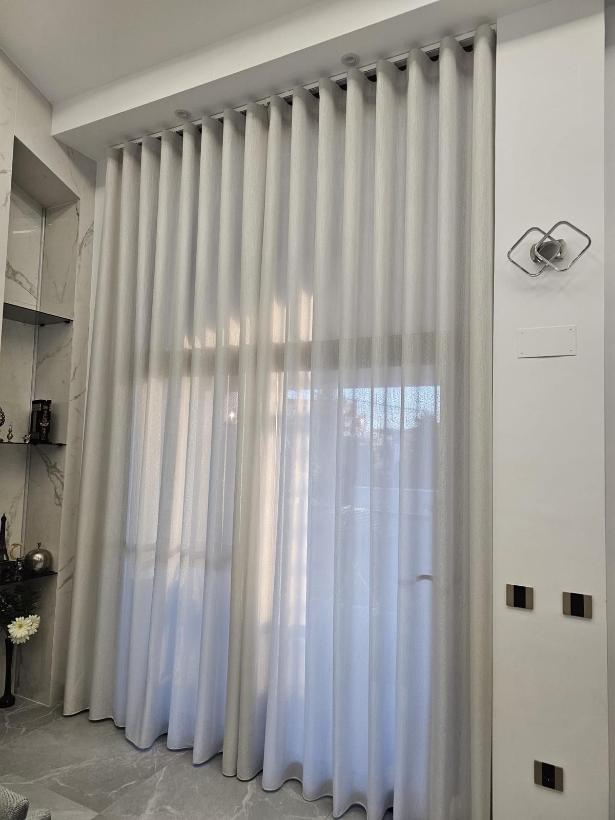 Unas cortinas blancas transparentes que llegan hasta el suelo cubren una ventana alta en una luminosa sala de estar.