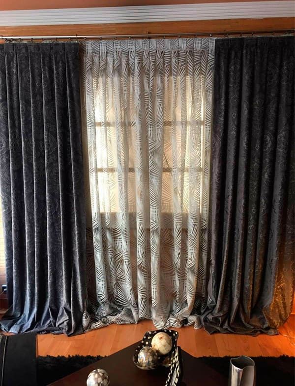 Cortinas gris oscuro enmarcan una ventana soleada con cortinas de encaje transparente y un cuenco decorativo sobre una mesita.