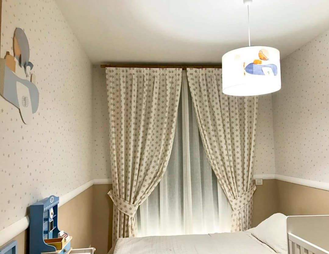Habitación infantil acogedora con paredes beige, cuna, ventana con cortinas y lámpara de techo colgante.
